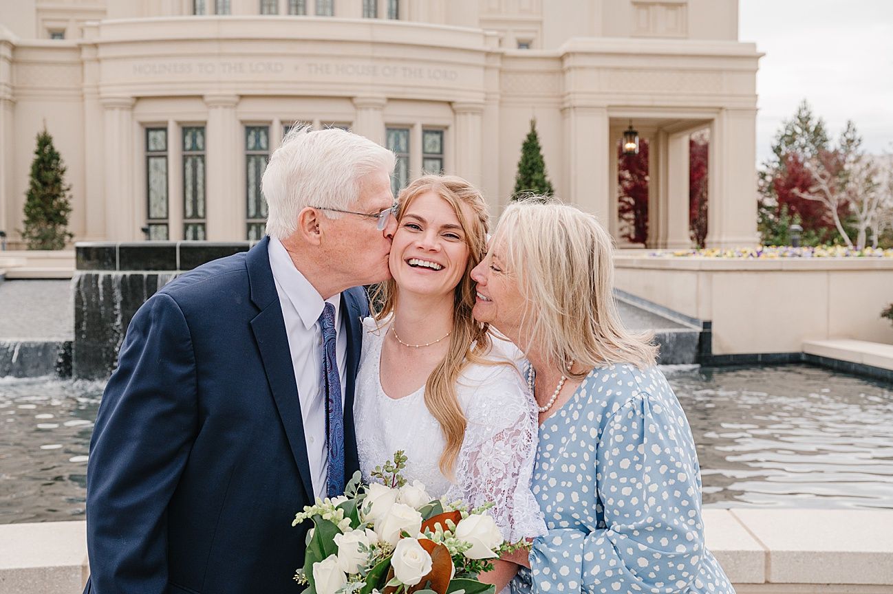 A bride in a white lace dress holds a bouquet while sharing a joyful moment outside an elegant building.