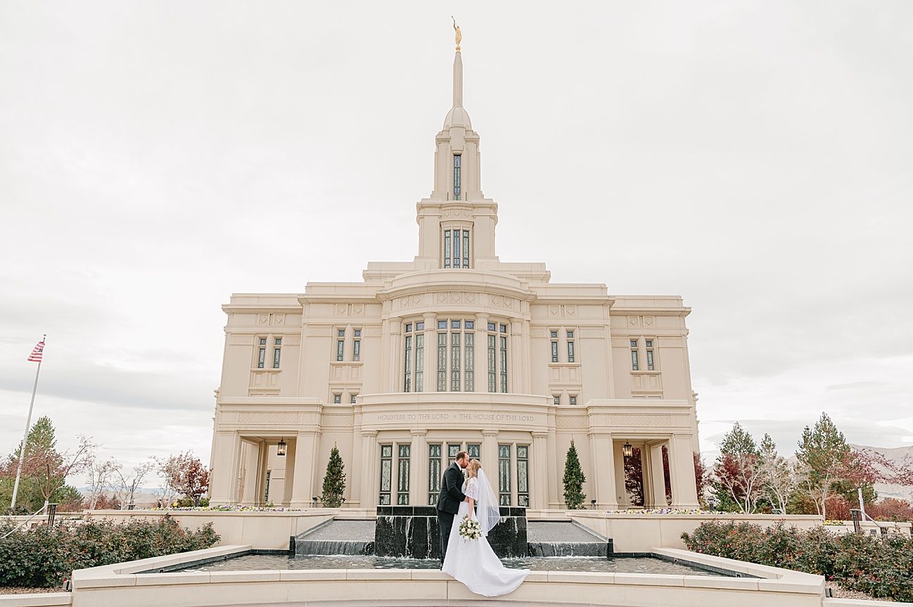 A couple stands in front of the elegant Payson Utah Temple, a majestic white building with a tall spire on a cloudy day.