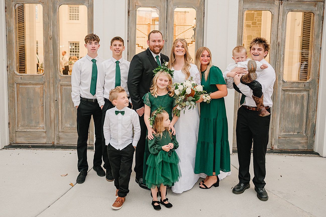 Wedding party lined up against white wall wearing emerald green dresses and black suits.