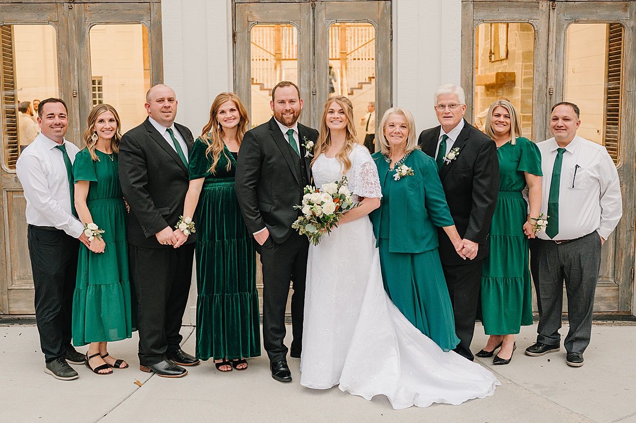 Wedding party members in emerald green and black formal wear pose in front of church windows.