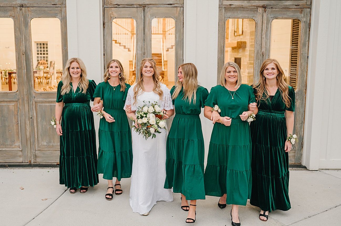 A group wearing coordinated emerald green dresses poses together against a light colored wall with windows.