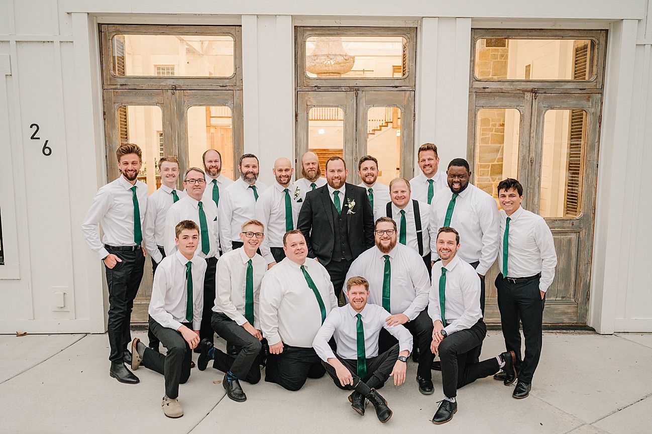 A panoramic wedding party photo showing groomsmen in white shirts and suspenders lined up against a white wall.