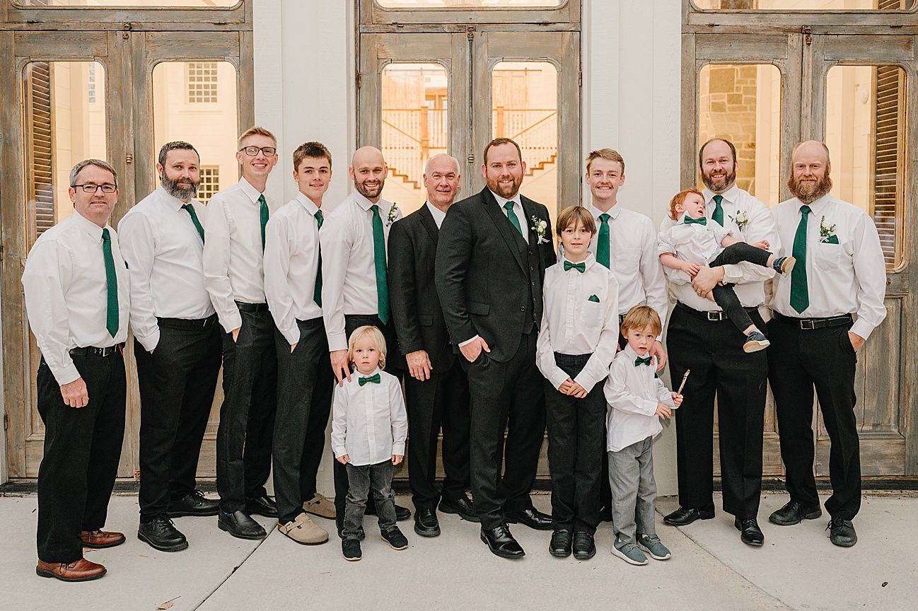 Wedding party members line up along stone building wall wearing formal attire.