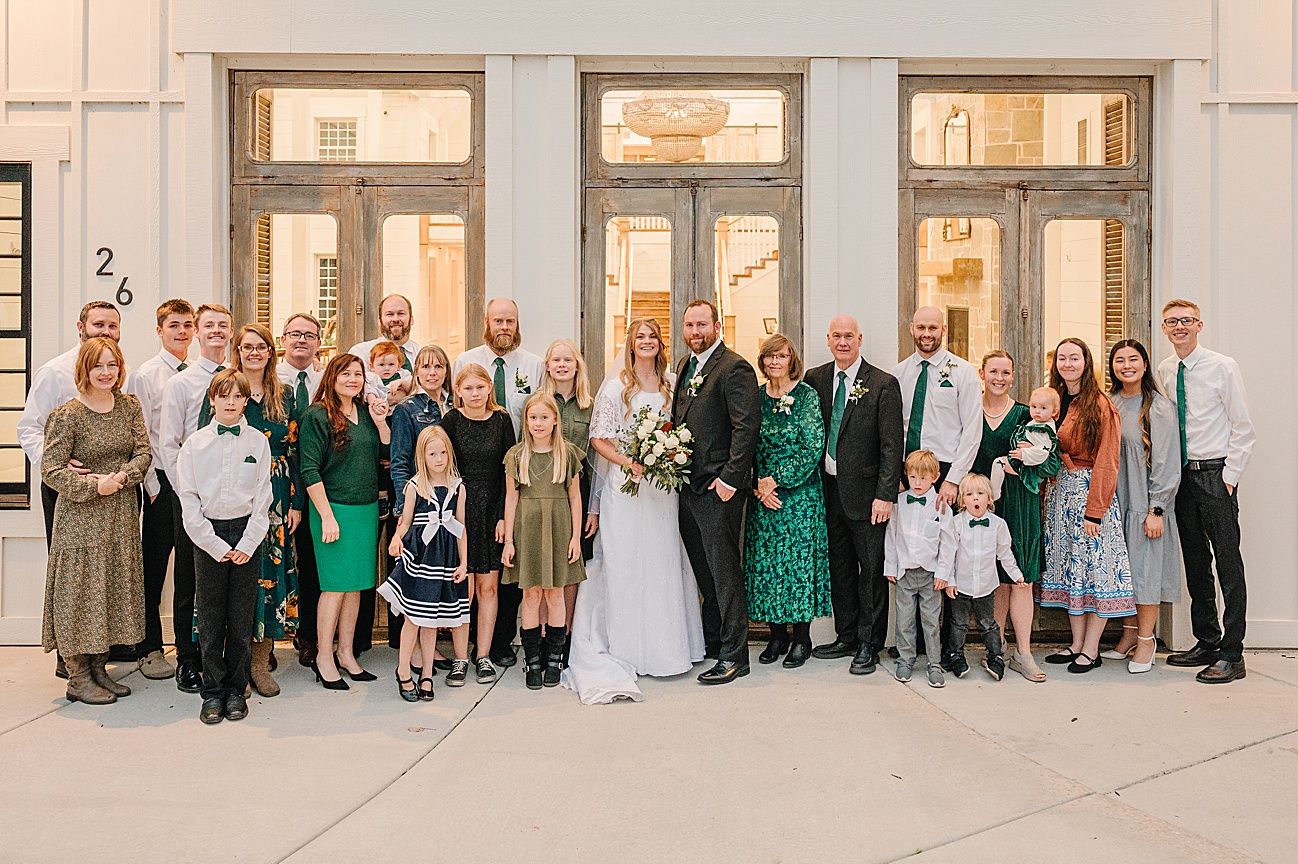 Wedding party and guests gathered in formal attire against a cream-colored wall with tall windows.