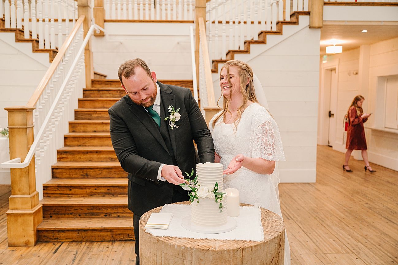 A couple in wedding attire cuts their cake together at the base of a wooden staircase during their reception.