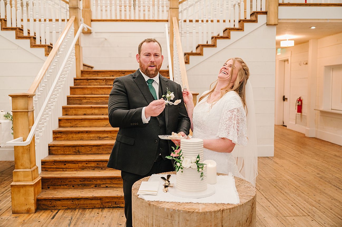 Newlyweds share a playful moment while cutting their wedding cake beside an elegant wooden staircase.