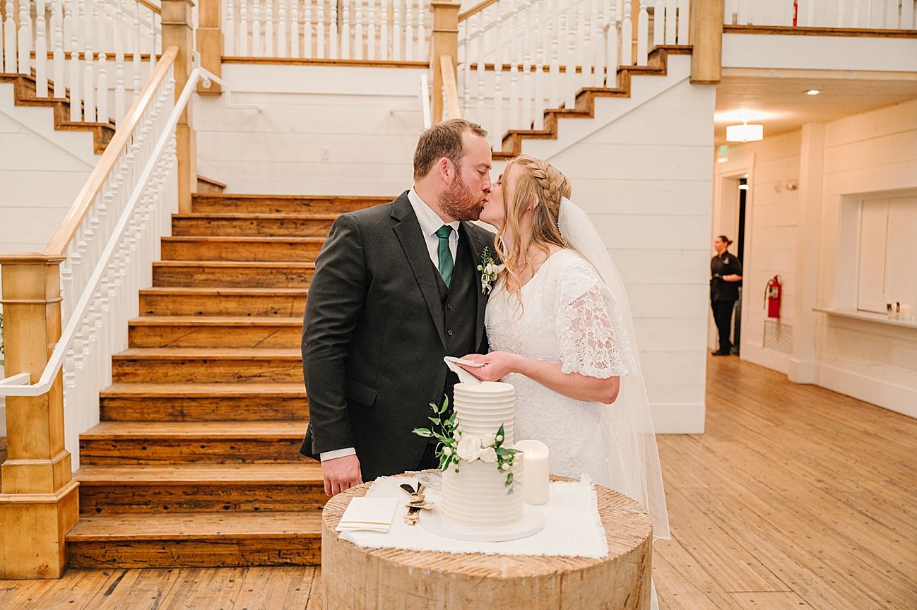 A couple shares a romantic moment by their wedding cake near an elegant wooden staircase in a bright indoor venue.