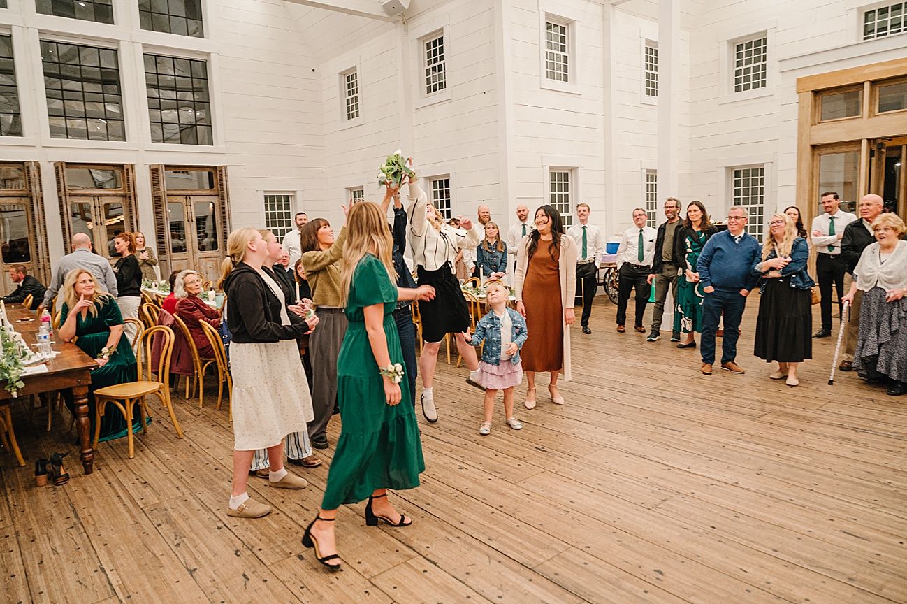 Wedding guests gather in a bright white indoor venue with rustic wooden floors and large windows.