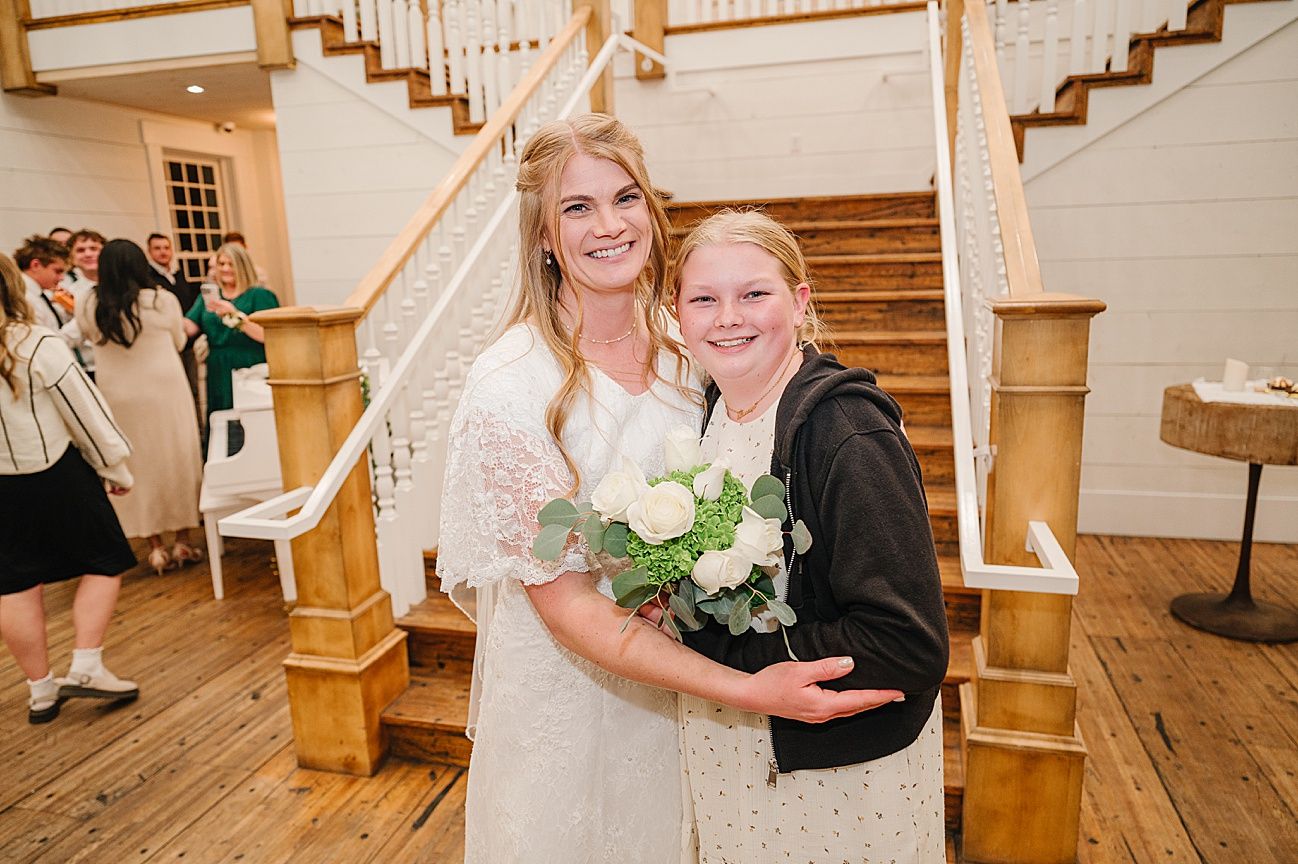 Two people share a joyful embrace in front of a wooden staircase at an indoor wedding venue.