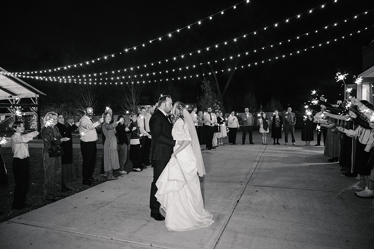 Wedding guests form a line under twinkling string lights at an outdoor evening reception.