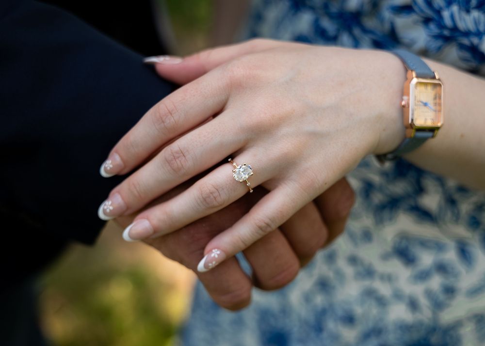 Elegant engagement ring and watch detail shot with neutral manicured nails against dark fabric.