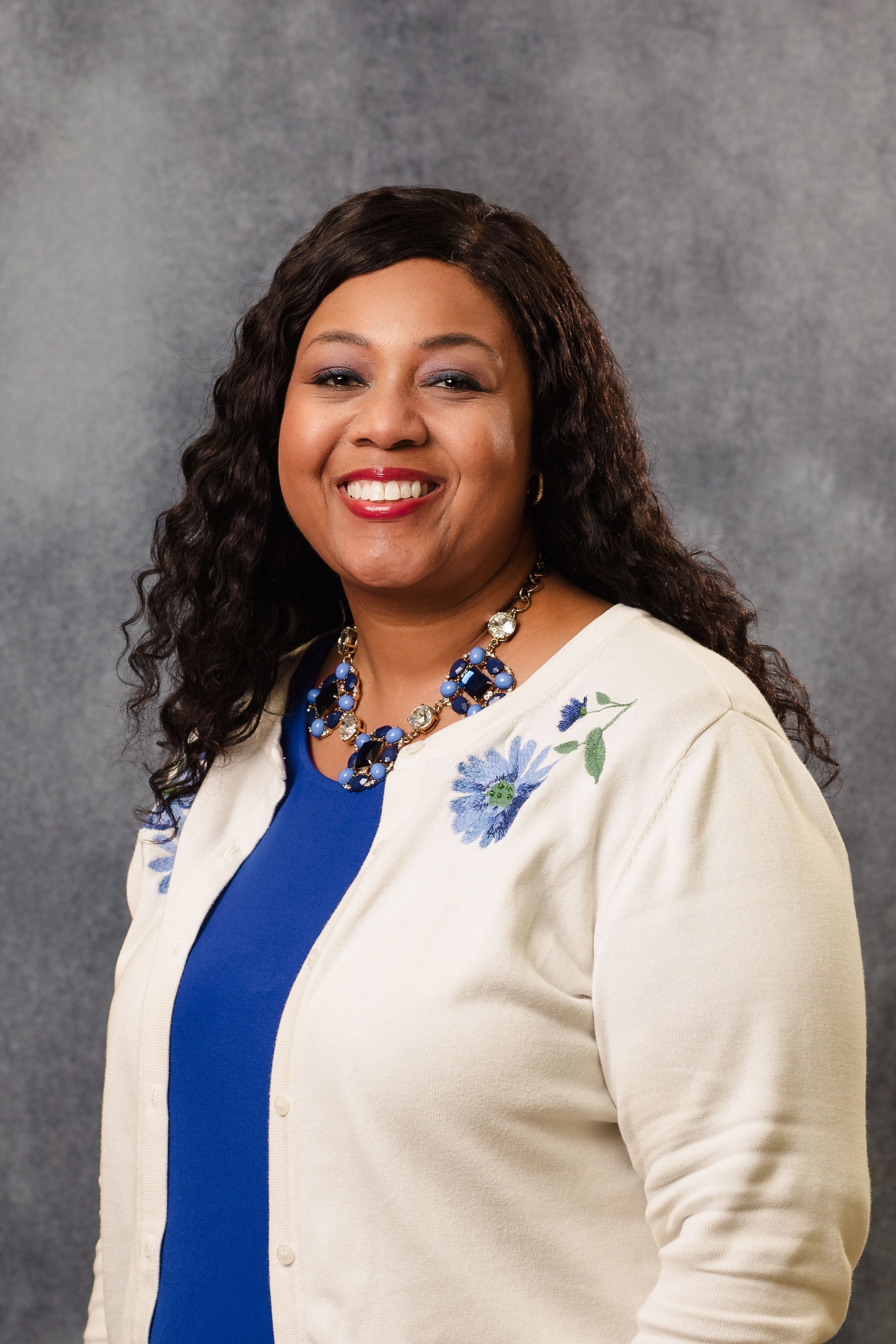 Professional headshot of person in white and blue outfit with floral details against grey background.
