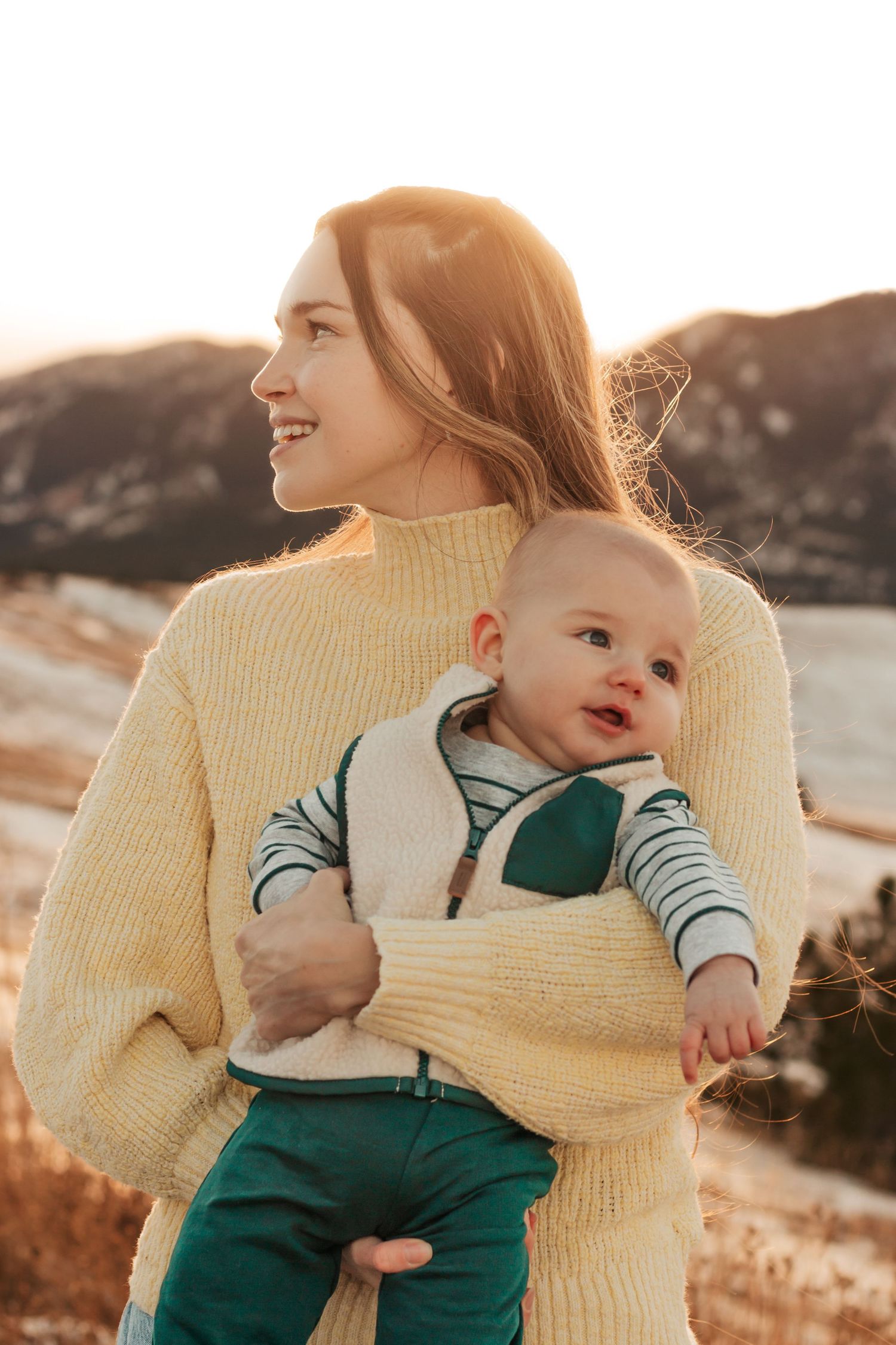 Parent in yellow sweater holds baby wearing green overalls during golden hour outdoor | Best Family Photo Location with Mountain View