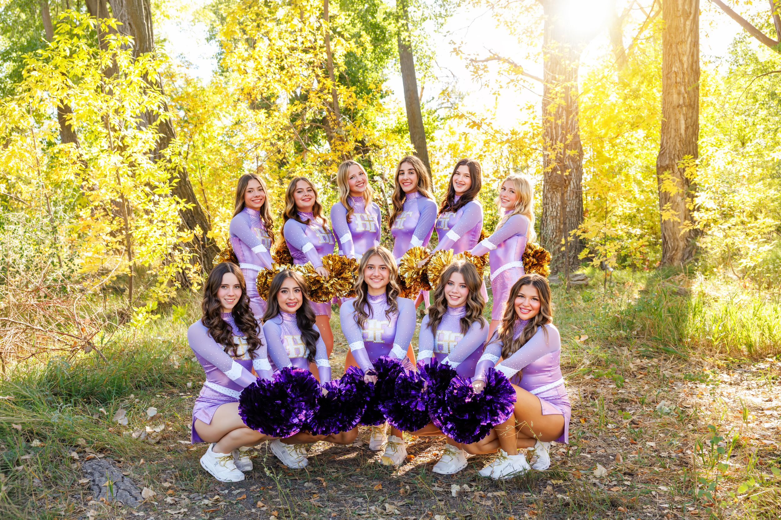 Cheerleaders in purple uniforms pose with gold and purple pom-poms outdoors among autumn trees in golden sunlight.