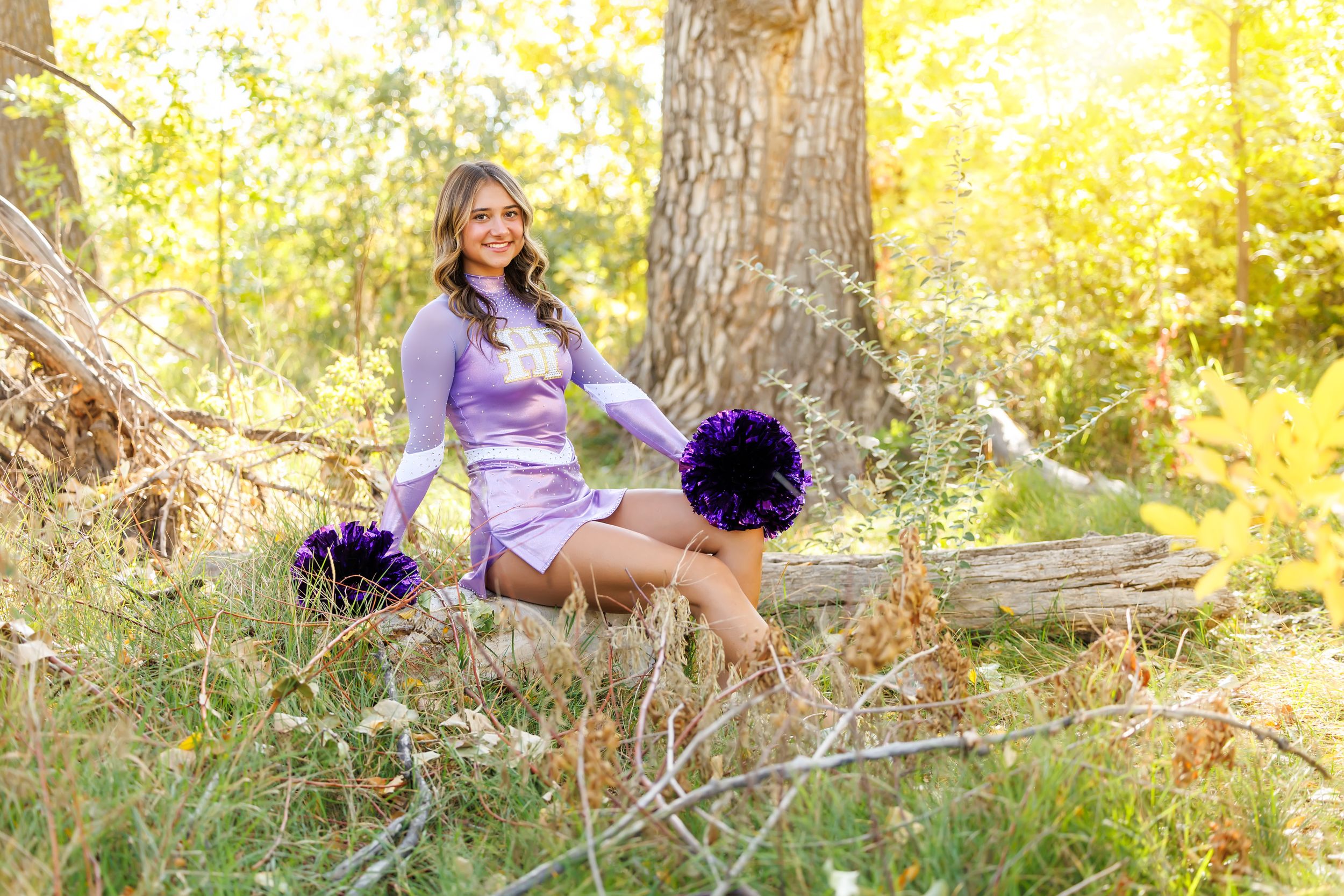 Cheerleader in purple uniform sitting on a fallen log in a sunlit forest, holding purple pom-poms.