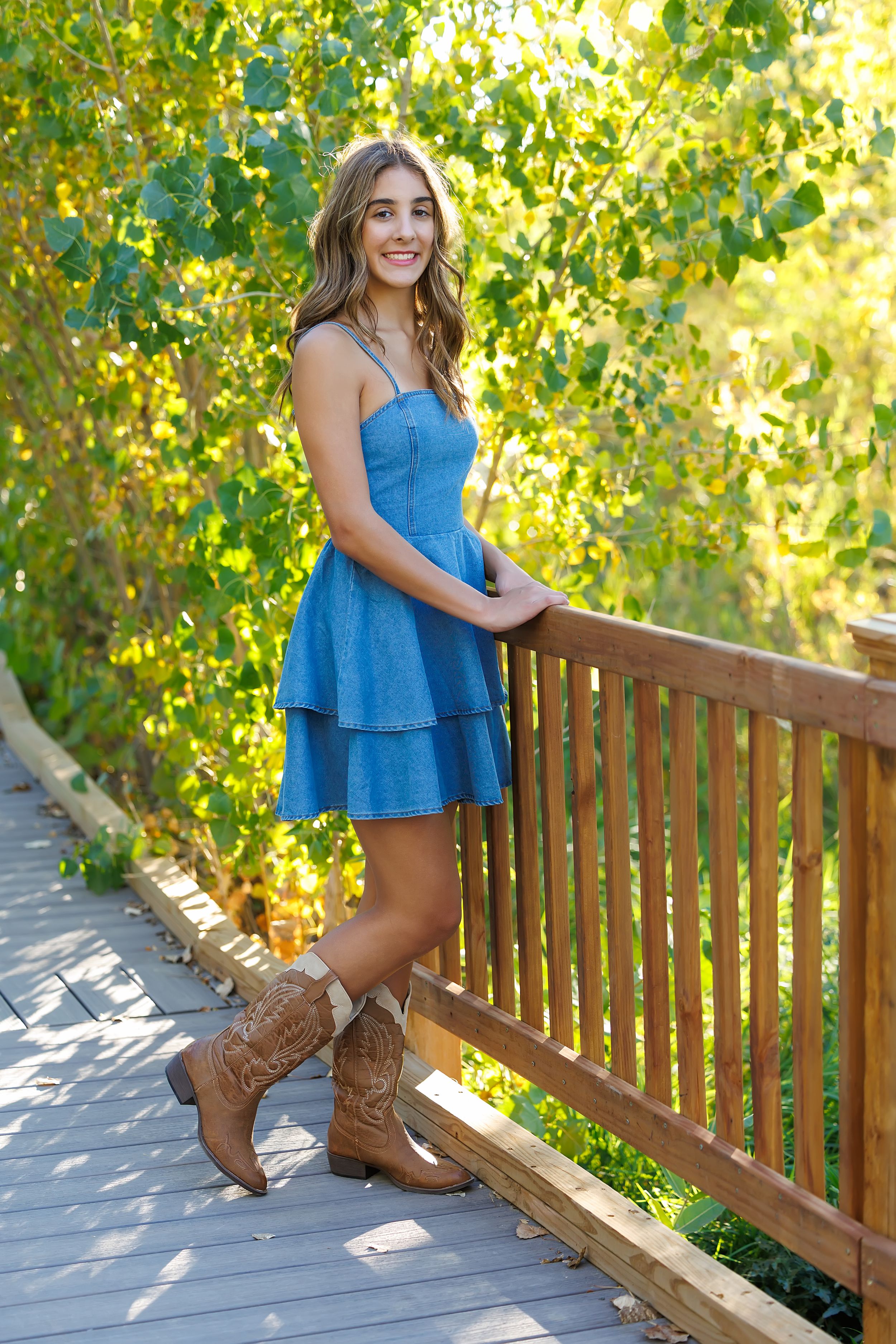 Young woman in denim dress and cowboy boots smiling on wooden bridge surrounded by lush green trees.