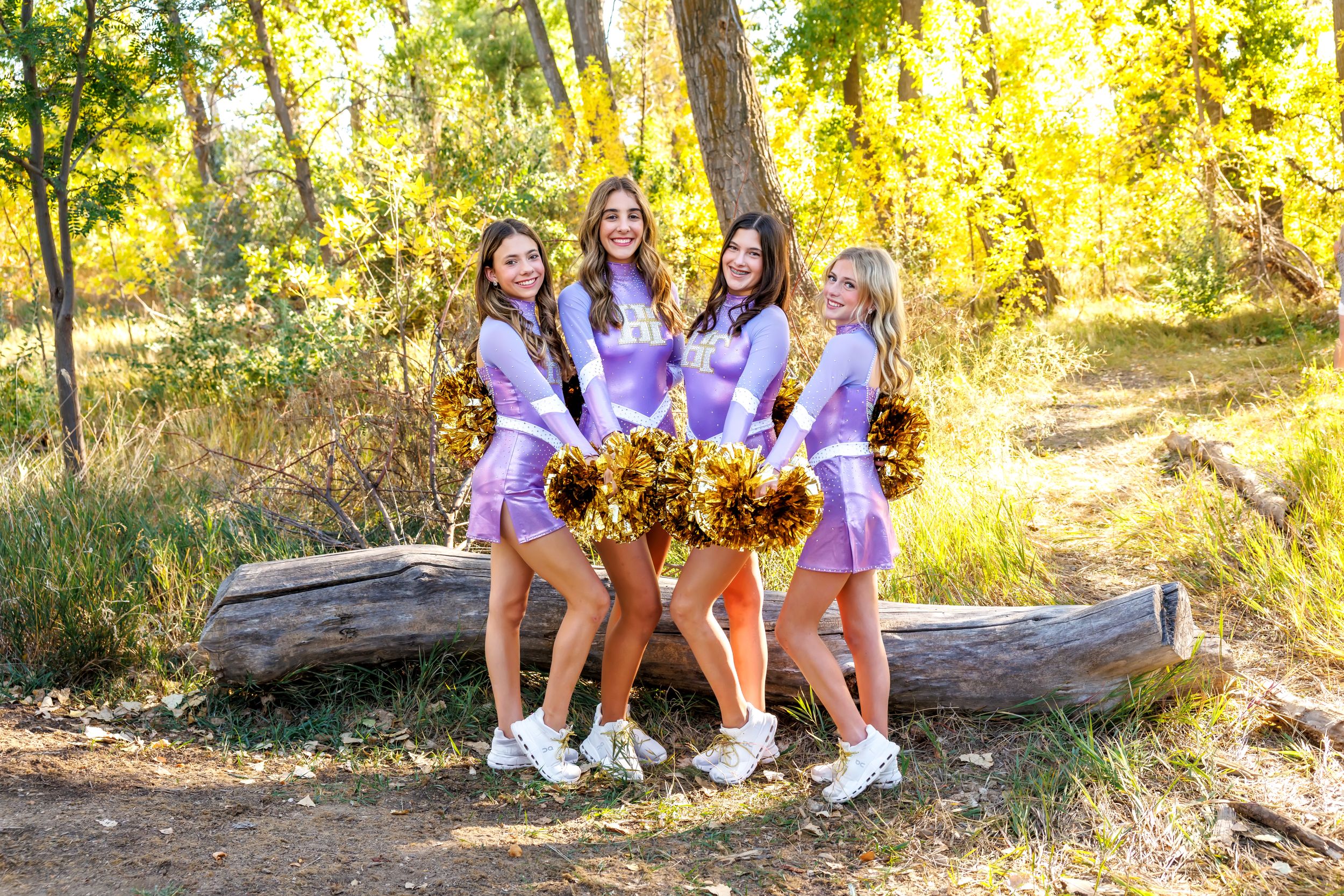 Four cheerleaders in purple uniforms holding gold pom-poms pose outdoors among autumn trees.