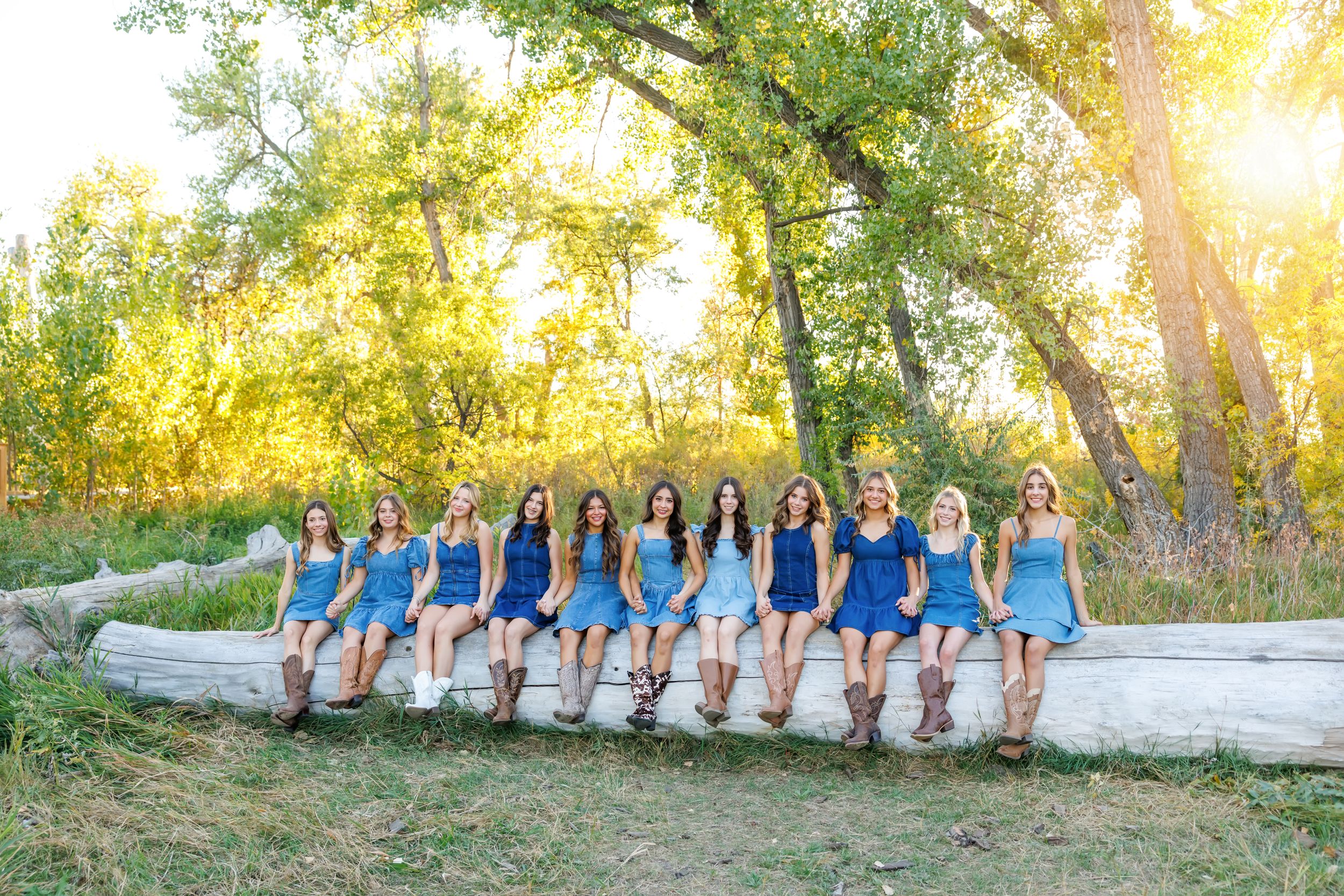 A group of women in blue dresses and cowboy boots sit on a log in a sunny outdoor setting.