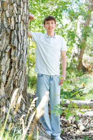 A smiling teenage boy in a white polo shirt and jeans stands beside a large tree trunk in a sunny forest.
