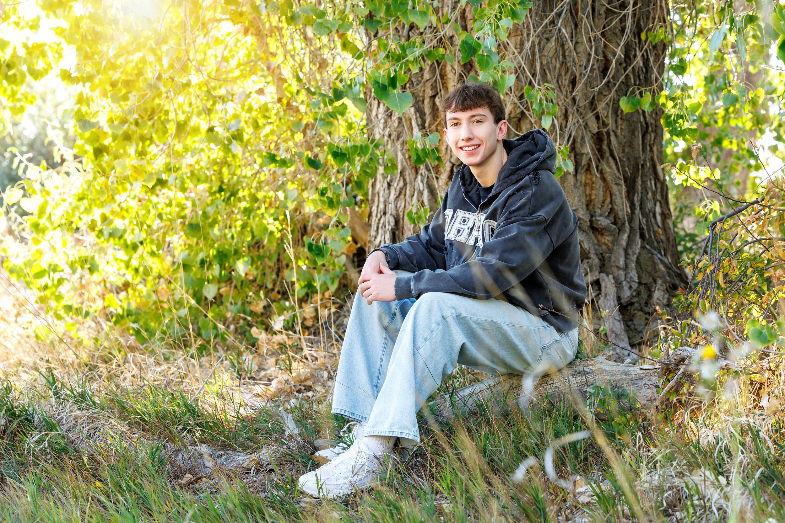 A teenage boy in a hoodie and jeans sits smiling under a large tree in a sunlit outdoor setting.