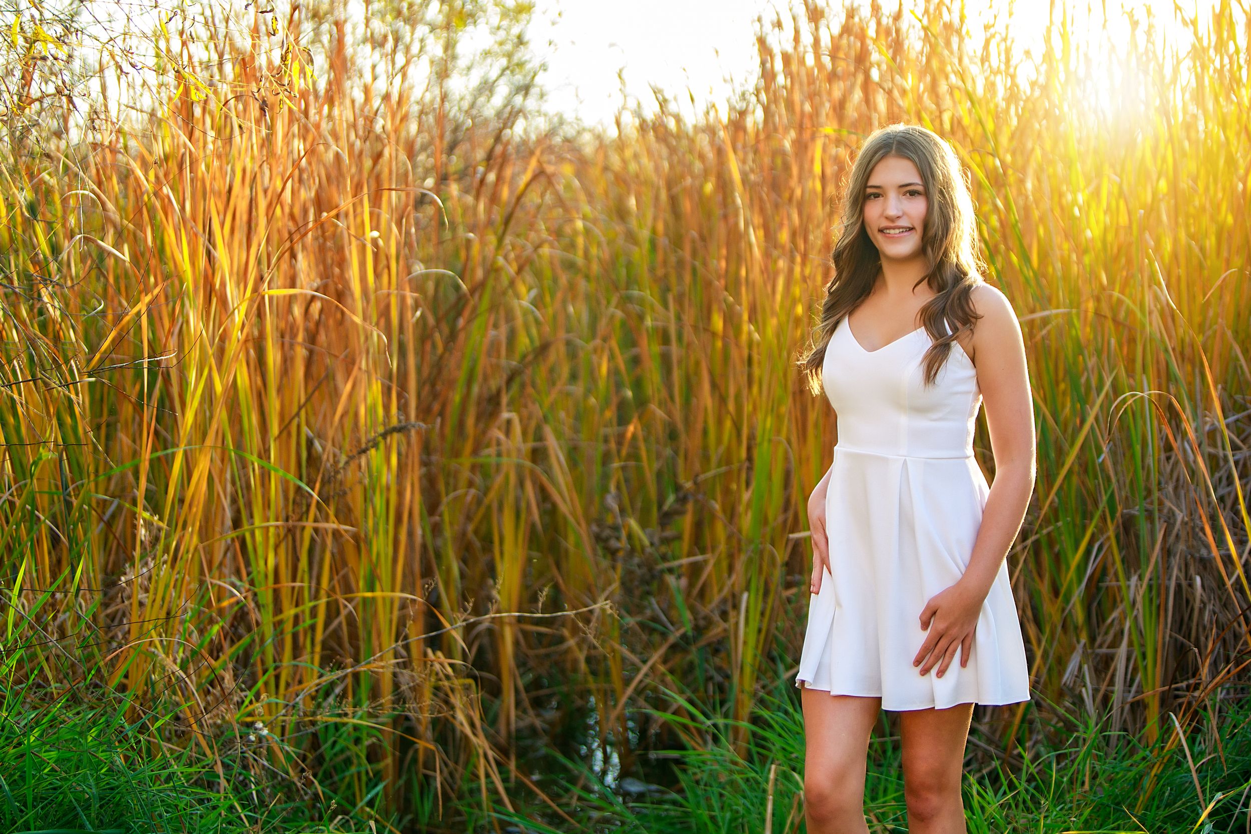 Woman in white dress poses in golden sunlit field of tall grass during sunset golden hour.