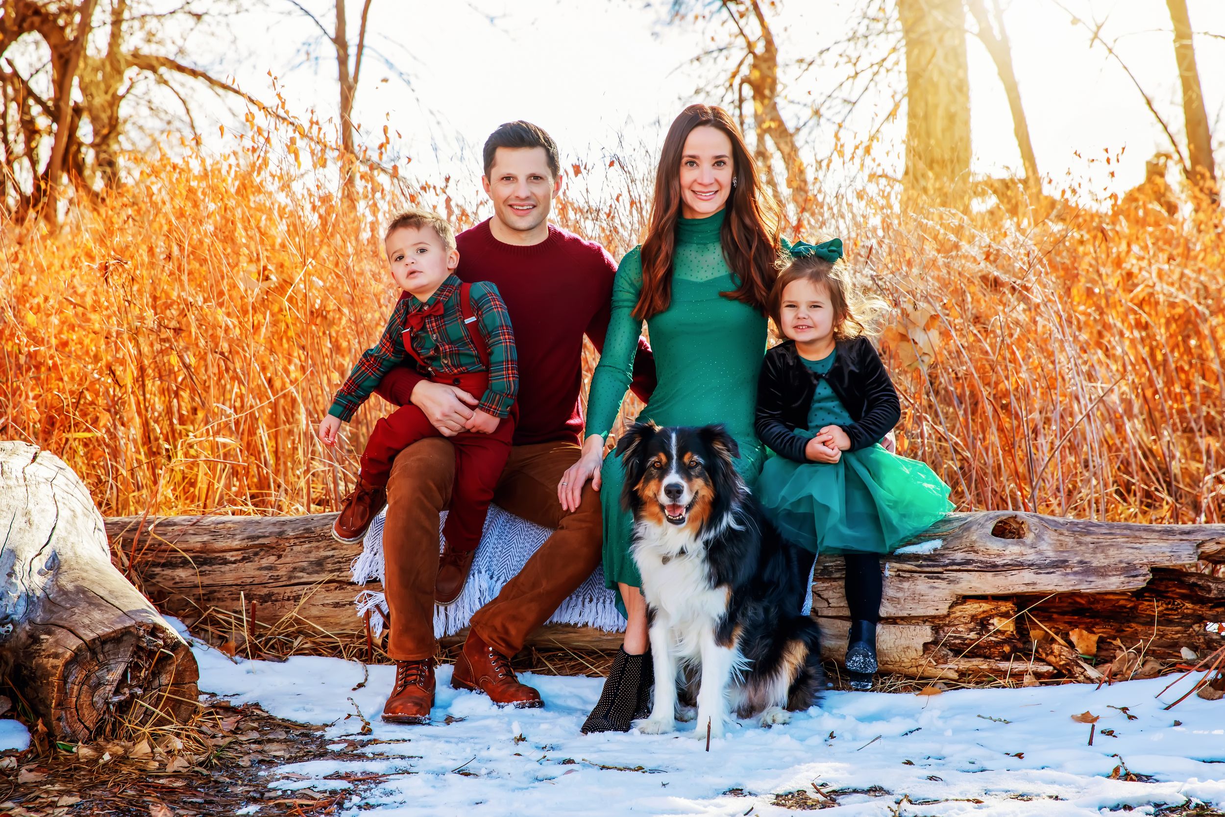 A family sits with their Australian Shepherd dog in a snowy winter setting during golden hour.