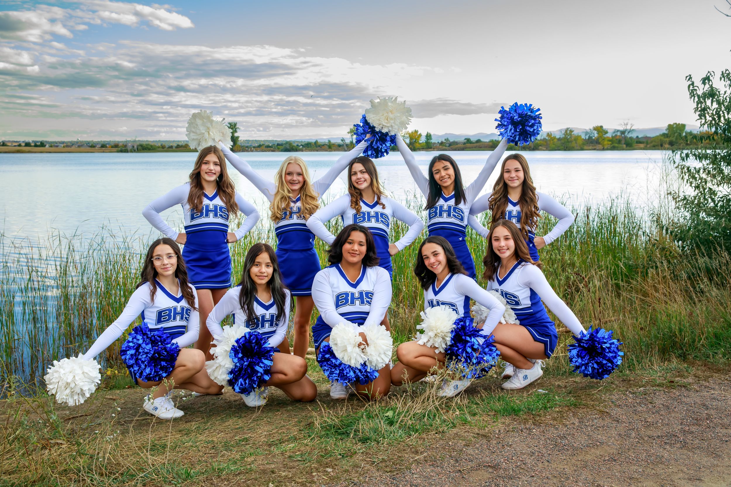 Cheerleaders in white and blue uniforms pose together on a lakeside path during a sunny summer day.