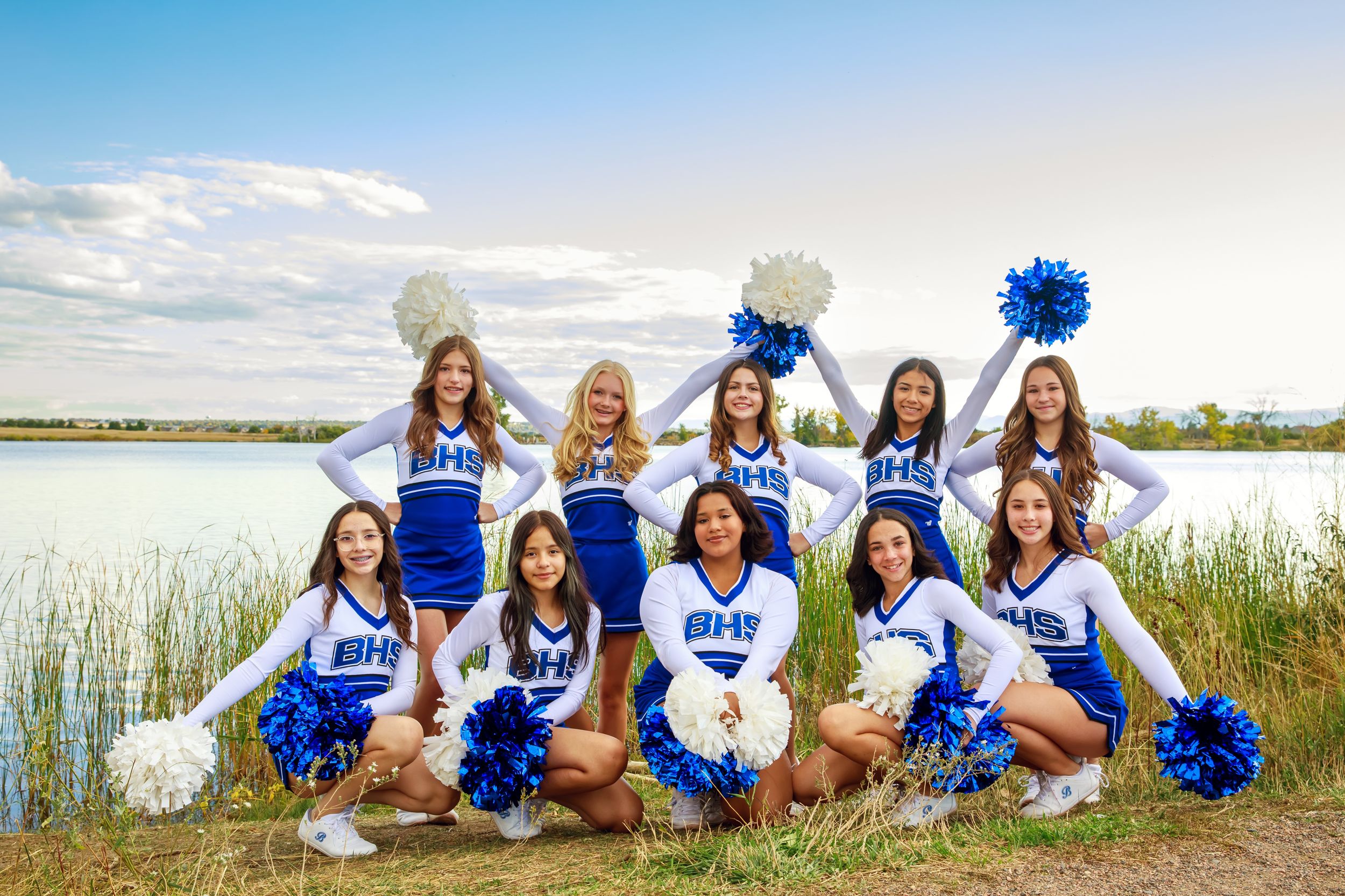 Cheerleaders in blue and white uniforms pose together on grass near a waterfront setting.
