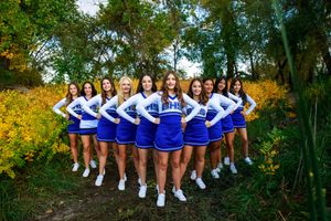 A cheerleading squad in blue and white uniforms poses together outdoors among yellow wildflowers and trees.