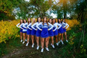 Cheerleading squad in blue and white uniforms poses in V-formation among yellow wildflowers and green trees.