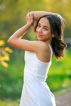 Woman in white tank top dancing joyfully outdoors with blurred green and yellow nature background.