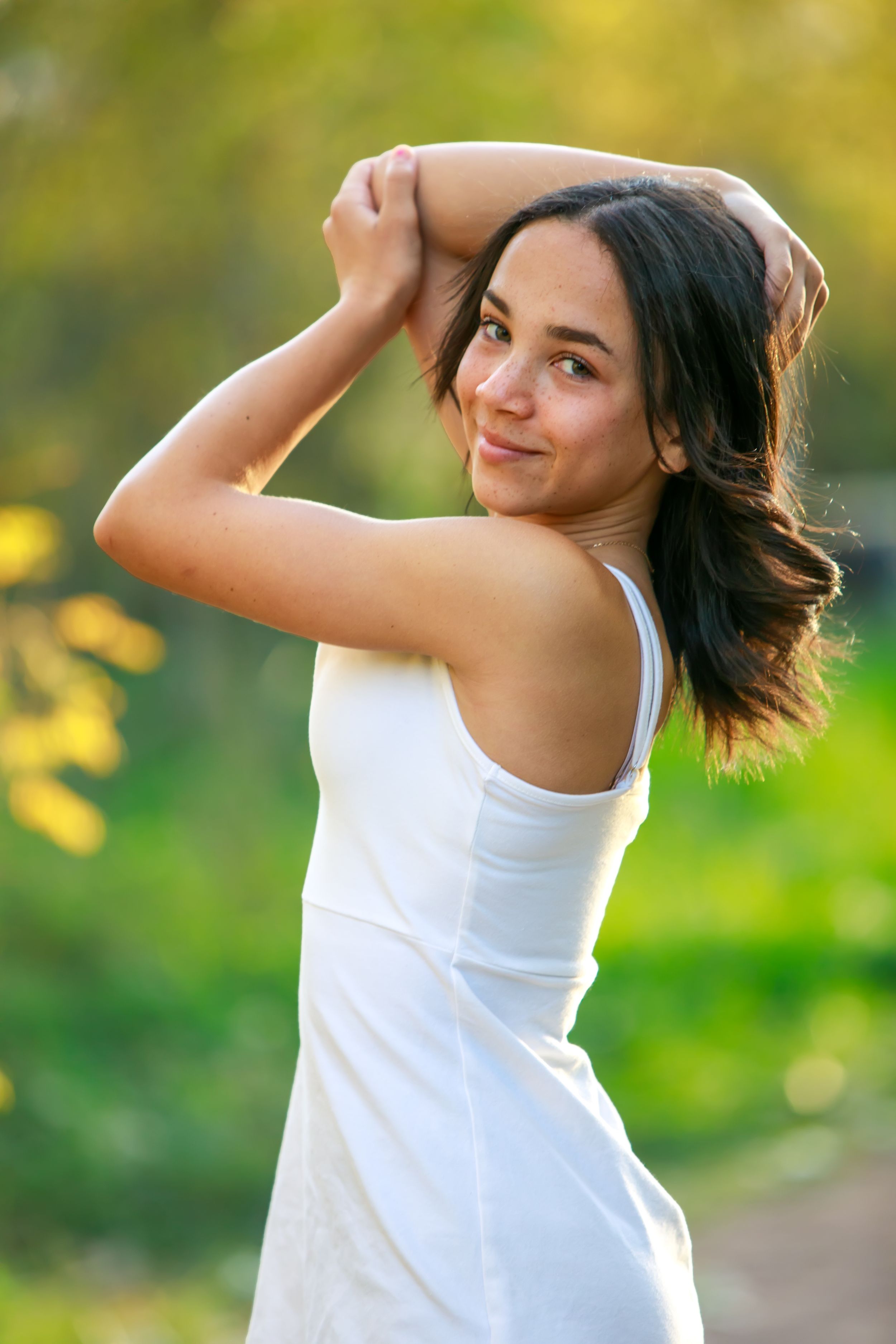 Woman in white tank top dancing joyfully outdoors with blurred green and yellow nature background.