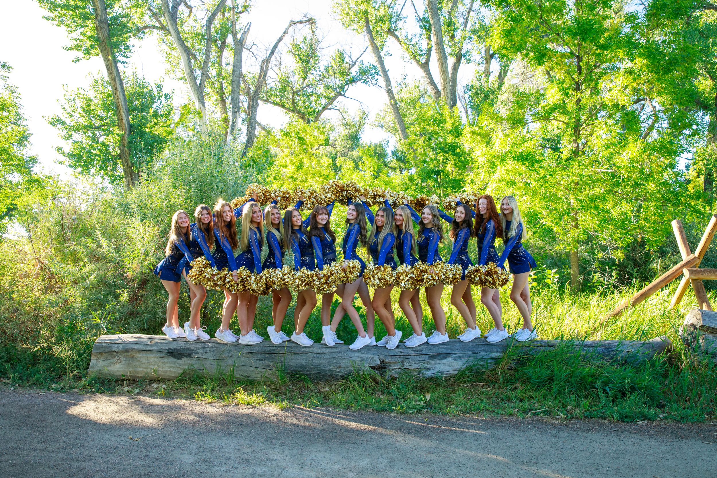 Large group of people in matching blue and yellow outfits pose together outdoors in front of green trees.