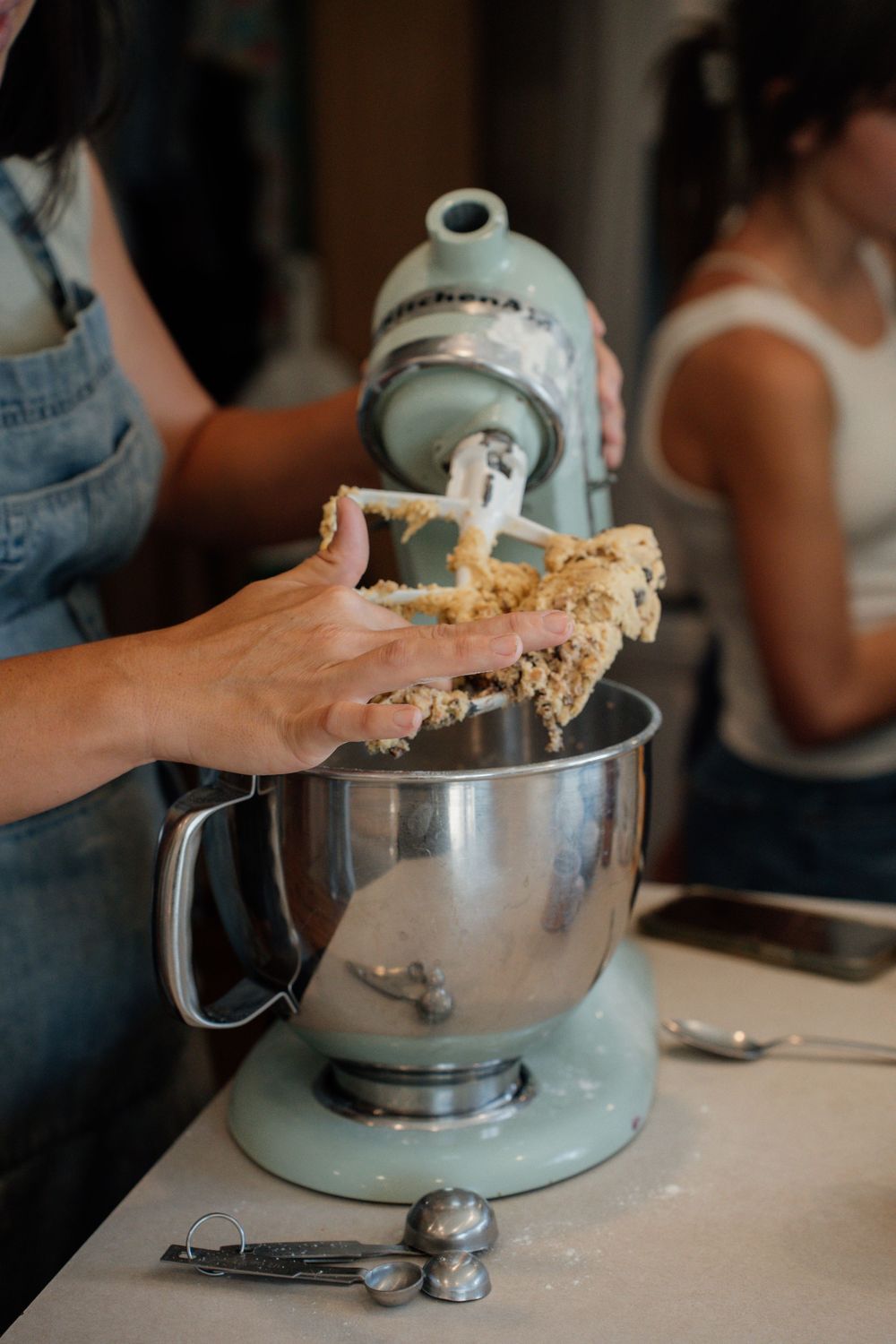 Hands adding dough mixture to a mint green stand mixer bowl on a white countertop during baking preparation.