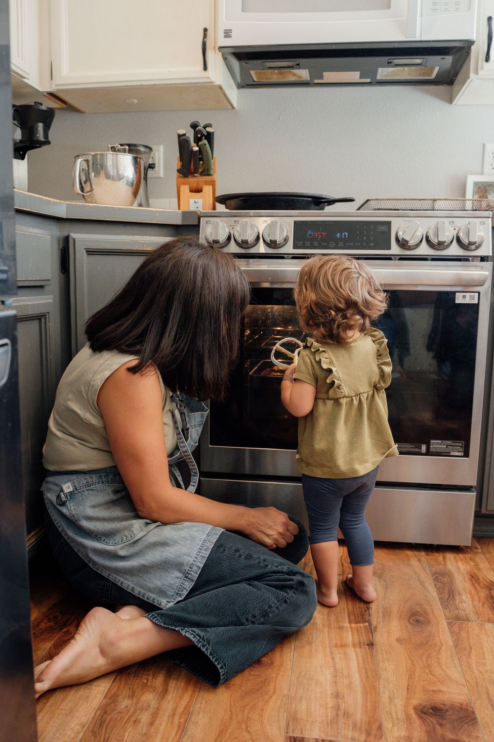 Mother and child looking inside an oven through glass door on hardwood floor.