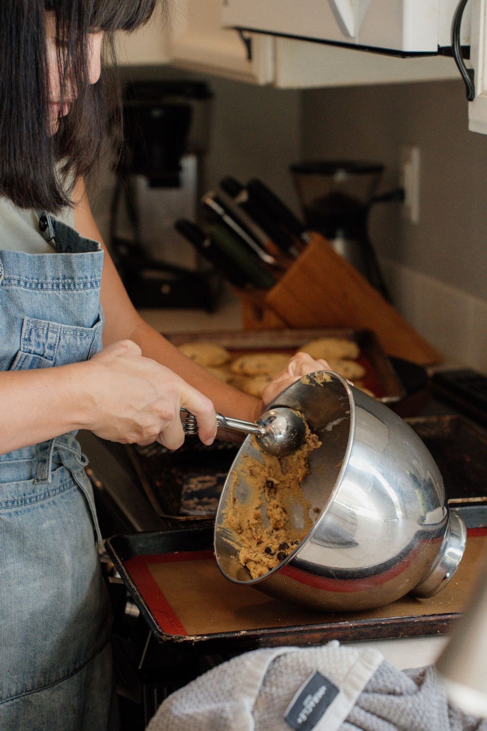 Person in denim overalls scraping cookie dough from a mixing bowl while baking in a kitchen.