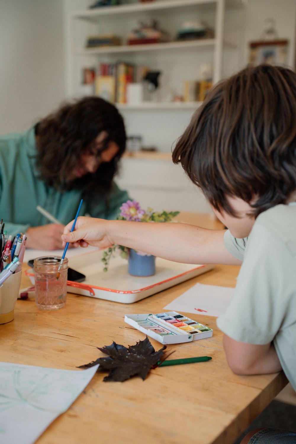 Student working on creative art project at wooden desk with watercolors and art supplies in bright classroom setting.