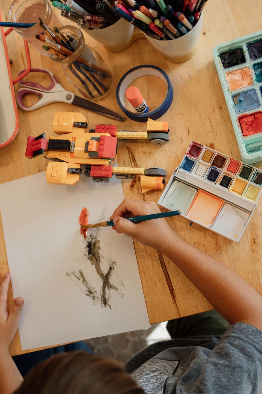 Overhead view of art supplies including watercolors, markers and paper with someone painting at a wooden desk.