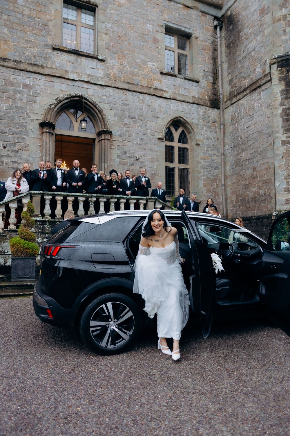 A wedding party exits a historic stone castle building next to a black luxury car parked on gravel.