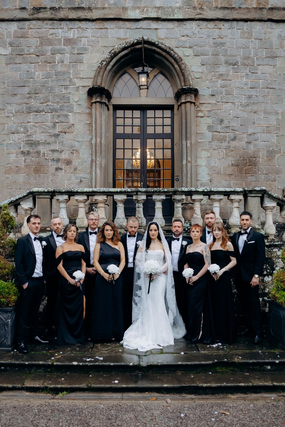 Wedding party in formal black and white attire poses together against stone castle wall at dusk.