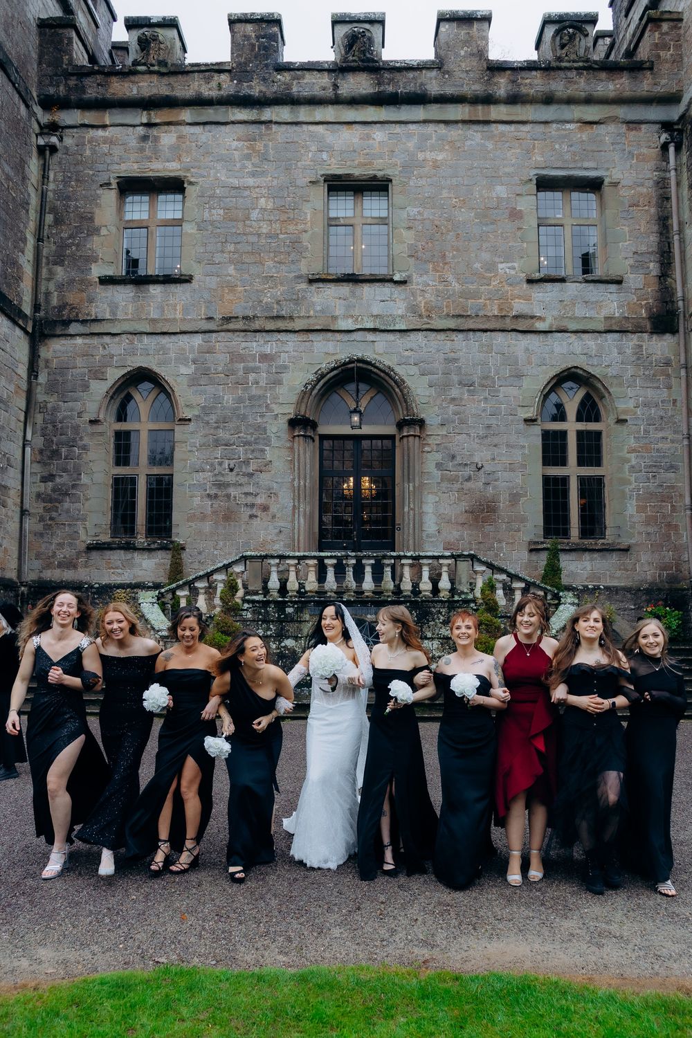 A wedding party poses together in front of a grand stone castle with Gothic windows and architecture.