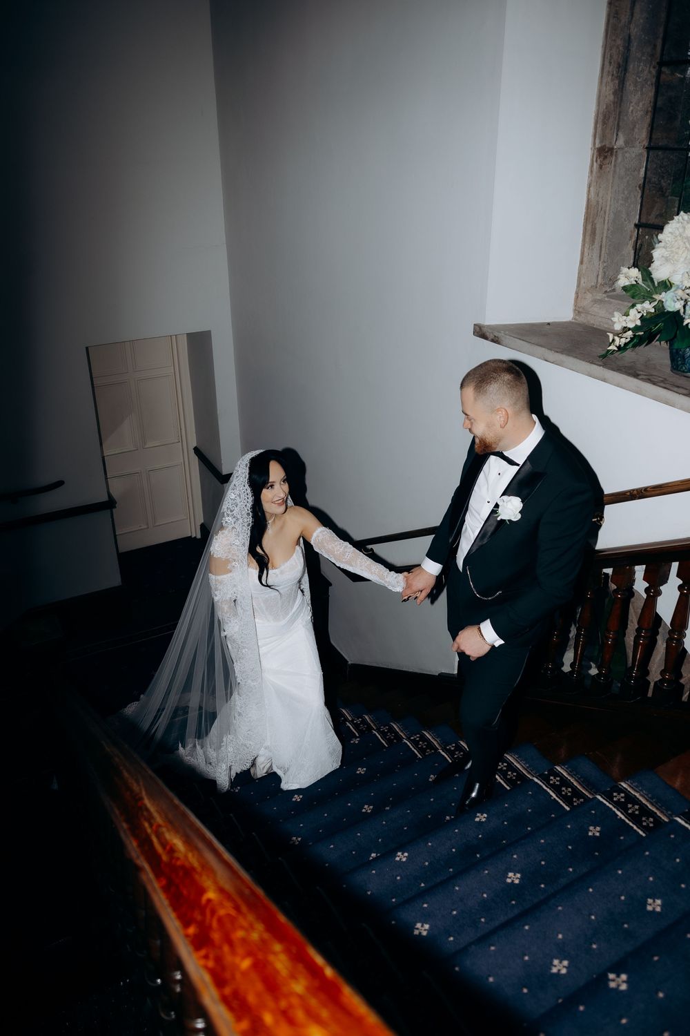 A couple in formal wedding attire hold hands while descending a dark staircase with star-patterned carpet.