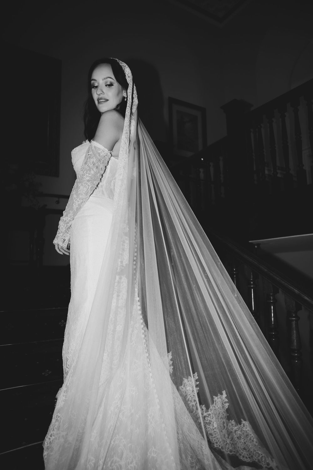 A bride in an elegant white lace wedding gown and flowing veil poses in dramatic black and white photography.