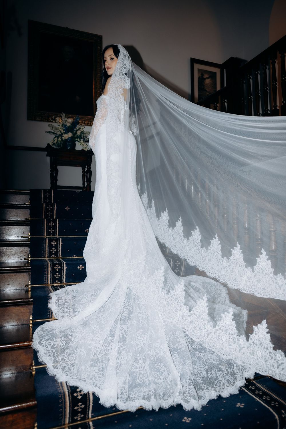 A flowing white wedding dress with intricate lace details and a dramatic cathedral-length veil cascades down wooden stairs.