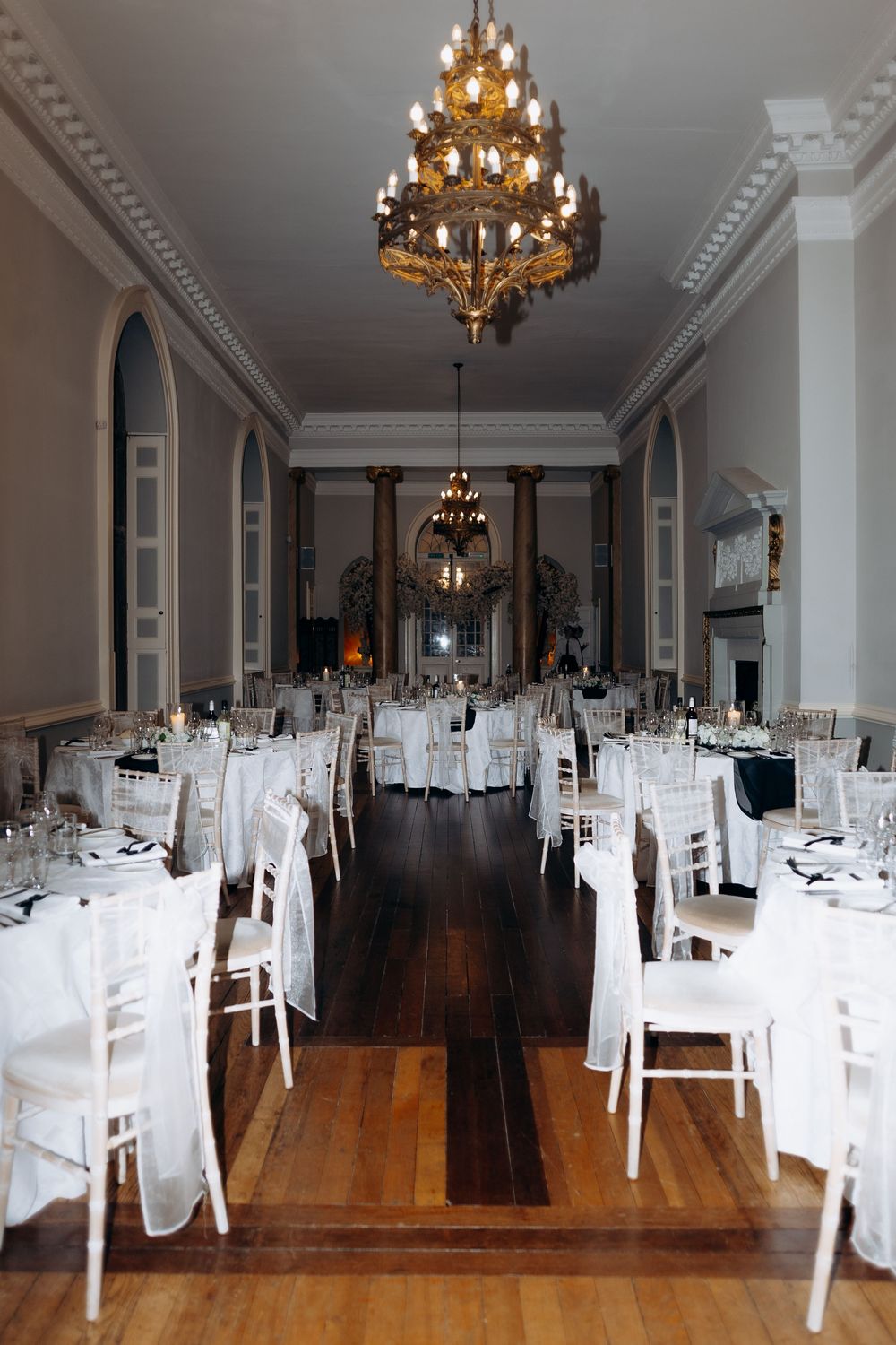 Elegant banquet hall with white chairs, hardwood floors, ornate chandeliers and crown molding in classic style.