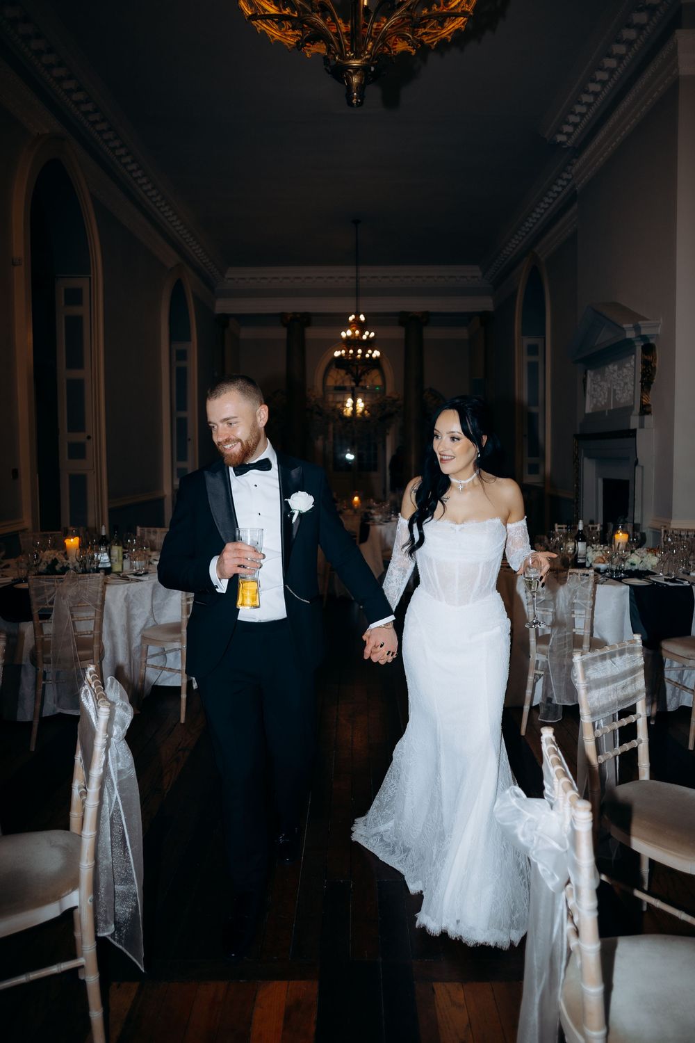 Wedding couple dances together in an elegant ballroom with ornate chandeliers and white chairs.