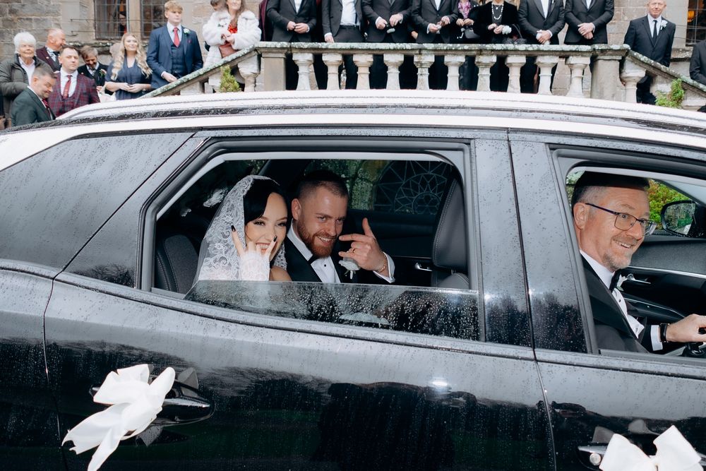 A black car decorated with white wedding ribbons and flowers drives away from a formal celebration.