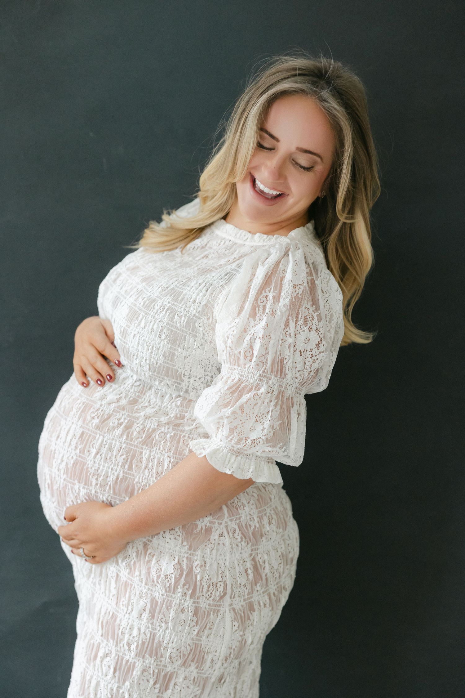 Expecting mother in white lace maternity dress smiles joyfully against dark background during pregnancy photoshoot.