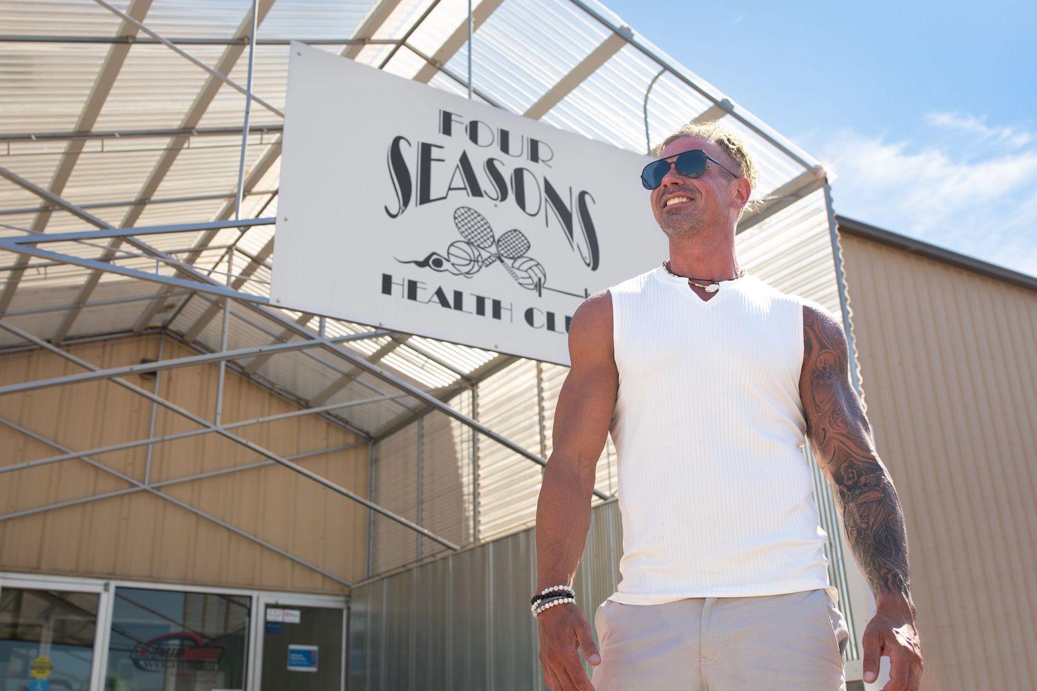 A Business Branding Session of a muscular personal trainer wearing a white sleeveless shirt standing in front of his gym on a sunny day.