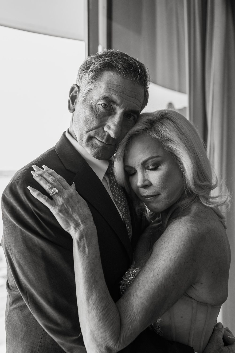 A romantic black and white portrait of a couple in formal attire embracing by a window.